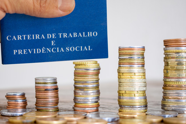 Foto de uma mão segurando uma carteira de trabalho e, abaixo, moedas empilhadas sobre a mesa